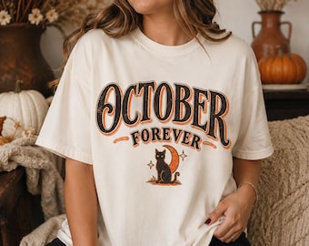 Könnte beinhalten: Ein cremefarbenes Kurzarm-T-Shirt mit dem Schriftzug "OCTOBER FOREVER" in schwarzer und orangefarbener Schrift. Unter dem Text befindet sich eine Grafik einer schwarzen Katze vor einem orangefarbenen Halbmond mit kleinen Sternen.