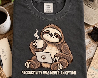 Könnte beinhalten: Ein anthrazitfarbenes T-Shirt mit einer Grafik eines Faultiers, das eine dampfende weiße Tasse hält und einen Laptop benutzt. Der Text unter dem Faultier lautet: "PRODUCTIVITY WAS NEVER AN OPTION". Am Kragen befindet sich ein Comfort Colors Etikett.