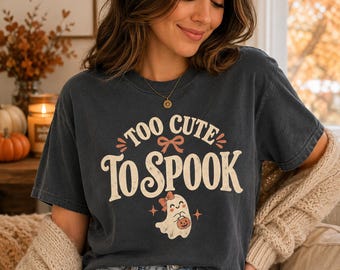 Könnte beinhalten: Ein anthrazitfarbenes Kurzarm-T-Shirt mit dem Schriftzug "TOO CUTE TO SPOOK" in cremefarbener Schrift und einer kleinen pfirsichfarbenen Schleife. Unter dem Text befindet sich eine Grafik eines weißen Geistes mit einer pfirsichfarbenen Schleife.