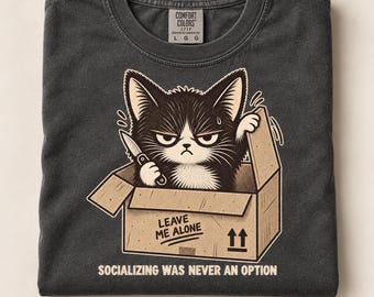 Könnte beinhalten: Ein anthrazitfarbenes T-Shirt mit einer Grafik eines mürrischen schwarz-weißen Kätzchens in einem Pappkarton, das ein kleines Messer hält. Auf dem Karton steht LEAVE ME ALONE und auf dem T-Shirt steht der Text SOCIALIZING WAS NEVER AN OPTION.