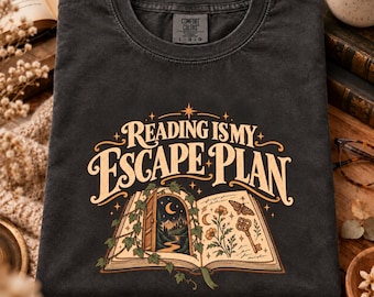 Könnte beinhalten: Ein anthrazitfarbenes T-Shirt mit Rundhalsausschnitt und dem Text Reading Is My Escape Plan in cremefarbener Schrift. Unter dem Text befindet sich eine Illustration eines offenen Buches mit Landschaft, Motte, Blumen und Schlüssel in Creme und Orange.