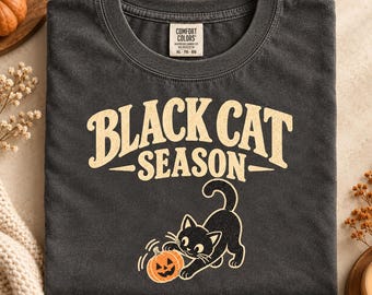 Könnte beinhalten: Ein anthrazitfarbenes Kurzarm-T-Shirt mit dem Text "BLACK CAT SEASON" in einer abgenutzten cremefarbenen Schrift. Unter dem Text befindet sich eine Grafik einer schwarzen Katze, die mit einem orangefarbenen Kürbis spielt. Comfort Colors Etikett.