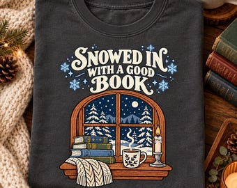 Könnte beinhalten: Ein dunkelgraues T-Shirt mit einer Grafik eines Fensters, das auf einen verschneiten Wald bei Nacht blickt. Der Text lautet SNOWED IN WITH A GOOD BOOK. Darunter sind drei gestapelte Bücher, eine dampfende Tasse, eine Kerze und eine Decke.