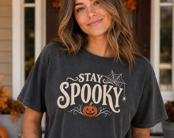 Könnte beinhalten: Ein anthrazitfarbenes Kurzarm-T-Shirt mit dem Schriftzug STAY SPOOKY in cremefarbener Schrift. Das Design zeigt ein Spinnennetz mit einer hängenden Spinne in der oberen rechten Ecke und einen orangefarbenen Kürbis am unteren Rand.