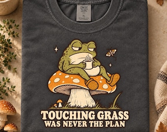 Könnte beinhalten: Ein anthrazitfarbenes T-Shirt mit einer Grafik eines grünen Frosches, der auf einem großen orangefarbenen Pilz mit weißen Punkten sitzt. Der Frosch hält eine weiße Tasse. Unter der Grafik steht der Text: TOUCHING GRASS WAS NEVER THE PLAN. Comfort Colors Etikett.