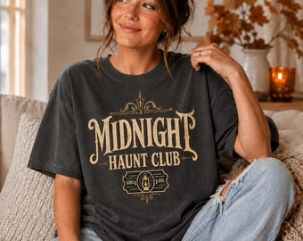 Könnte beinhalten: Ein anthrazitfarbenes Kurzarm-T-Shirt mit einem goldenen Grafikdruck, der Midnight Haunt Club liest. Das Design umfasst verzierte Schnörkel, ein Laternen-Symbol und einen rechteckigen, ticketartigen Rahmen mit dem Text Admit One.
