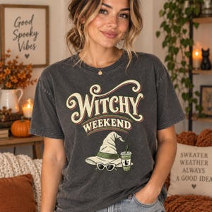 Può includere: Una t-shirt a maniche corte grigio antracite con la scritta "WITCHY WEEKEND" in carattere color crema e rosso. Sotto il testo c'è la grafica di un cappello da strega verde chiaro, occhiali da sole bianchi cat-eye e un bicchiere di caffè freddo.