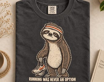 Könnte beinhalten: Ein anthrazitfarbenes T-Shirt mit einer Grafik eines Faultiers, das ein orange-weißes Stirnband trägt und einen Laufschuh hält. Der Text unter dem Faultier lautet "RUNNING WAS NEVER AN OPTION". Das Shirt hat ein Comfort Colors Etikett am Kragen.