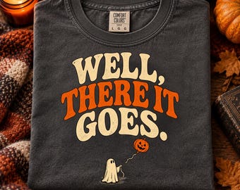 Könnte beinhalten: Ein anthrazitfarbenes T-Shirt mit dem Text WELL, THERE IT GOES in großen, cremefarbenen und orangefarbenen Retro-Buchstaben. Unter dem Text befindet sich eine kleine Illustration eines weißen Geistes, der eine Schnur zu einem orangefarbenen Kürbisballon hält.