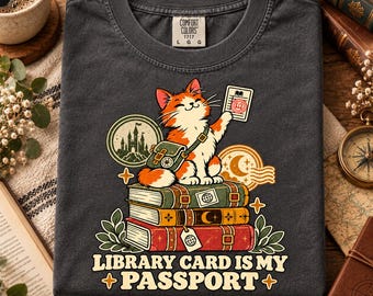 Könnte beinhalten: Ein anthrazitfarbenes T-Shirt mit einer Grafik einer orange-weißen Katze, die auf einem Stapel aus drei Büchern sitzt. Die Katze trägt eine grüne Umhängetasche und hält einen Bibliotheksausweis. Unter den Büchern steht der Text "LIBRARY CARD IS MY PASSPORT."