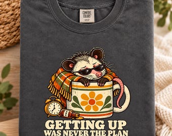 Könnte beinhalten: Ein anthrazitfarbenes T-Shirt mit einer Grafik eines Opossums mit Sonnenbrille, eingewickelt in eine karierte Decke, das in einer Blumentasse ruht. Daneben stehen ein Wecker und eine Flasche. Der Text lautet: GETTING UP WAS NEVER THE PLAN.