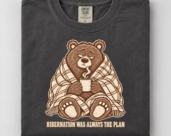 Könnte beinhalten: Ein anthrazitfarbenes T-Shirt mit einer Grafik eines braunen Bären, der in eine karierte Decke eingewickelt ist und eine dampfende Tasse hält. Der Text unter dem Bären lautet, HIBERNATION WAS ALWAYS THE PLAN. Das Comfort Colors Etikett ist am Kragen.