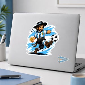 K&ouml;nnte beinhalten: Ein Aufkleber mit einer Cartoon-Illustration eines Fu&szlig;ballspielers in einem Argentinien-Trikot, der einen Fu&szlig;ball tritt. Der Spieler tr&auml;gt einen schwarzen Hut und die Nummer 10. Der Hintergrund hat blaue und wei&szlig;e Akzente, und ein kleines Argentinien-Wimpel ist sichtbar.