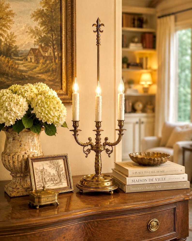 May include: A brass candelabra with three lit candles sits on a wooden table, next to a vase of white flowers, a framed picture, and a small decorative box. A stack of books and a decorative bowl are also on the table. A painting hangs on the wall.