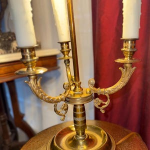 May include: A brass candelabra with three candle holders and a central light. The candelabra has ornate, curved arms and a round base. The candles are white and have a dripped wax appearance. The background includes a red curtain and a table.