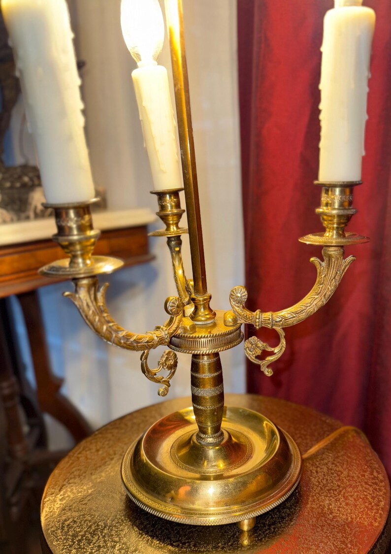 May include: A brass candelabra with three candle holders and a central light. The candelabra has ornate, curved arms and a round base. The candles are white and have a dripped wax appearance. The background includes a red curtain and a table.