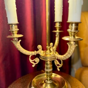 May include: A vintage brass candelabra with three candle holders. The candelabra has a detailed design with curved arms and decorative accents. The candles are white and have a dripped wax appearance. The candelabra is on a round table.