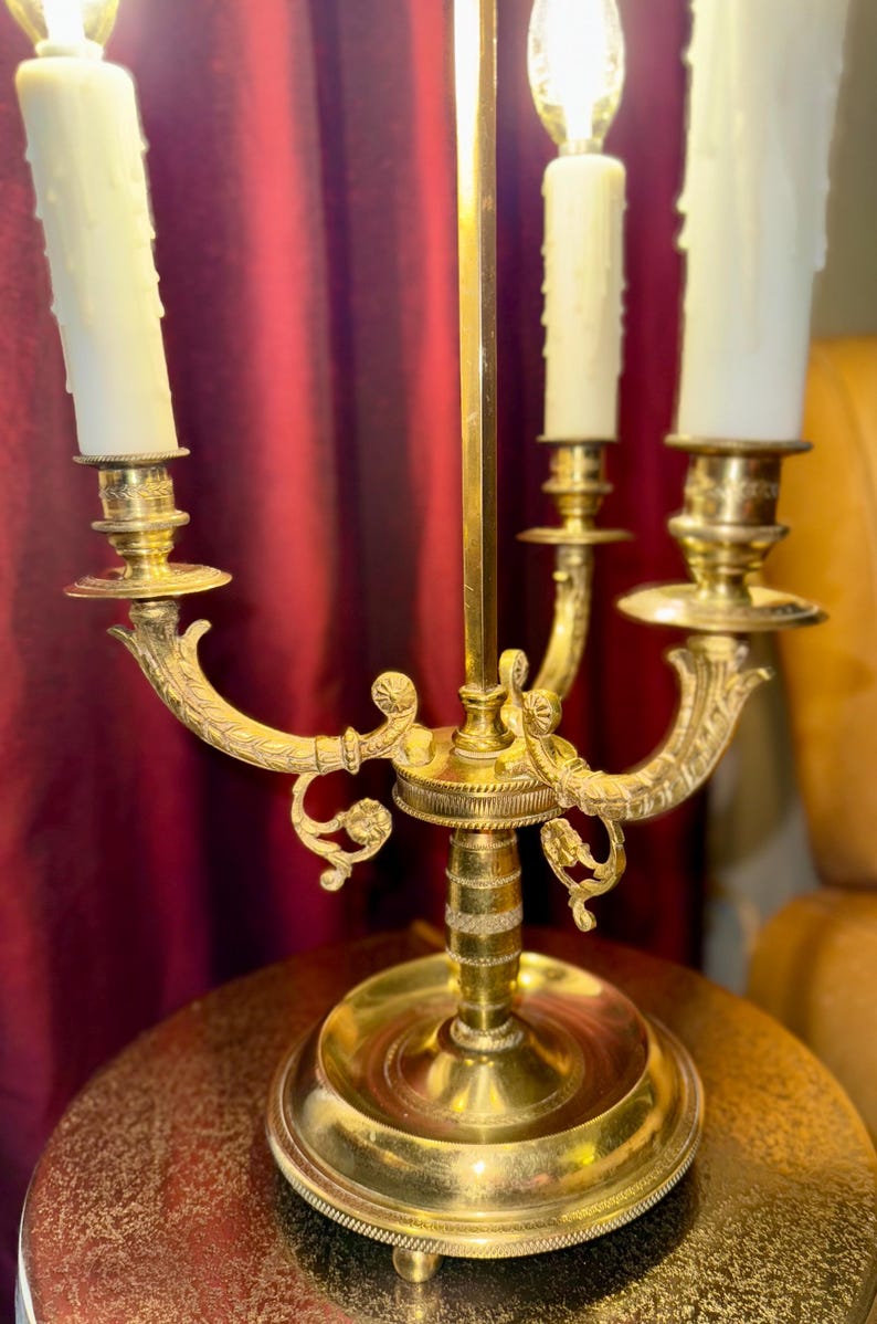 May include: A vintage brass candelabra with three candle holders. The candelabra has a detailed design with curved arms and decorative accents. The candles are white and have a dripped wax appearance. The candelabra is on a round table.