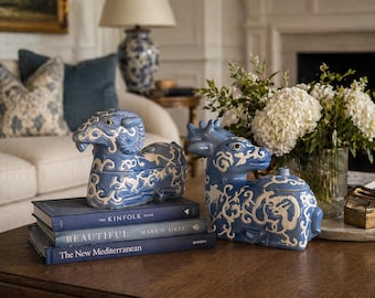May include: Two blue ceramic animal figurines, a ram and a deer, with white ornate detailing. They sit on a stack of blue books on a wooden coffee table. A vase of white flowers and a sofa are in the background.