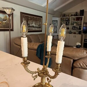 May include: A brass candelabra-style lamp with three faux candle lights. The lamp has a decorative base and arms, with a fleur-de-lis finial. The bulbs are clear and the candle sleeves are white.