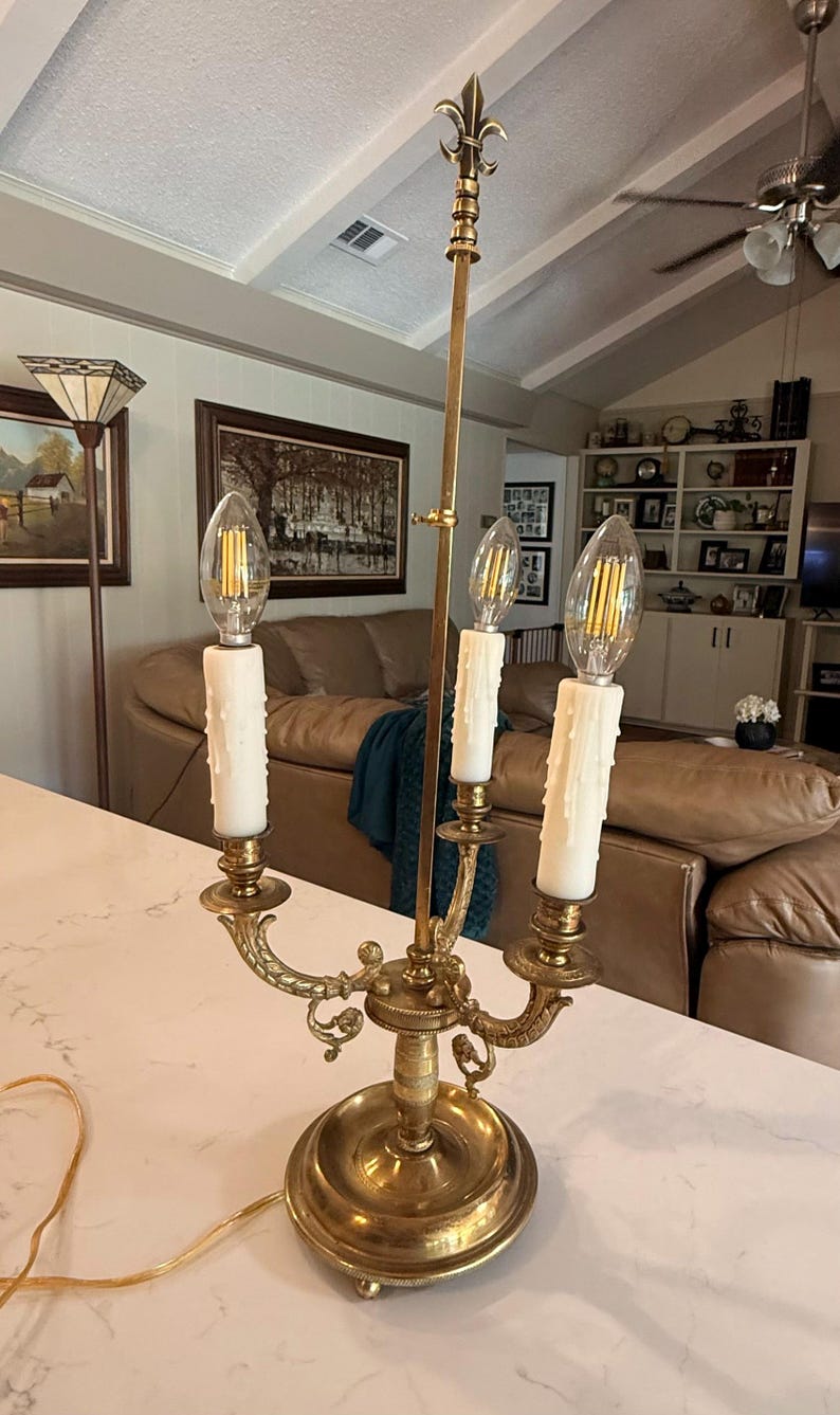 May include: A brass candelabra-style lamp with three faux candle lights. The lamp has a decorative base and arms, with a fleur-de-lis finial. The bulbs are clear and the candle sleeves are white.