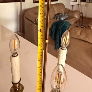 May include: A brass-colored, three-light candelabra-style lamp with a fleur-de-lis finial. The lamp has three faux-candle light bulbs. The height of the lamp is measured at approximately 19 inches.