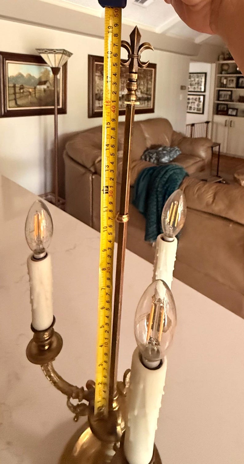 May include: A brass-colored, three-light candelabra-style lamp with a fleur-de-lis finial. The lamp has three faux-candle light bulbs. The height of the lamp is measured at approximately 19 inches.