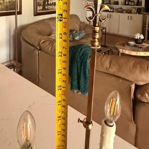 May include: A brass and gold-toned floor lamp with a fleur-de-lis design at the top. The lamp is approximately 60 inches tall and features two candle-shaped light bulbs.