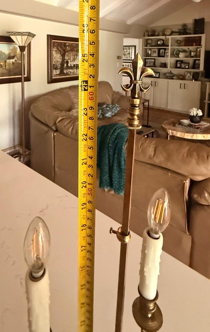 May include: A brass and gold-toned floor lamp with a fleur-de-lis design at the top. The lamp is approximately 60 inches tall and features two candle-shaped light bulbs.