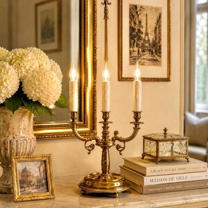 May include: A gold-toned candelabra with three candle holders, set on a marble-topped table. The candelabra is flanked by framed artwork, a decorative glass box, and a vase of white flowers. The scene evokes a classic, elegant aesthetic.
