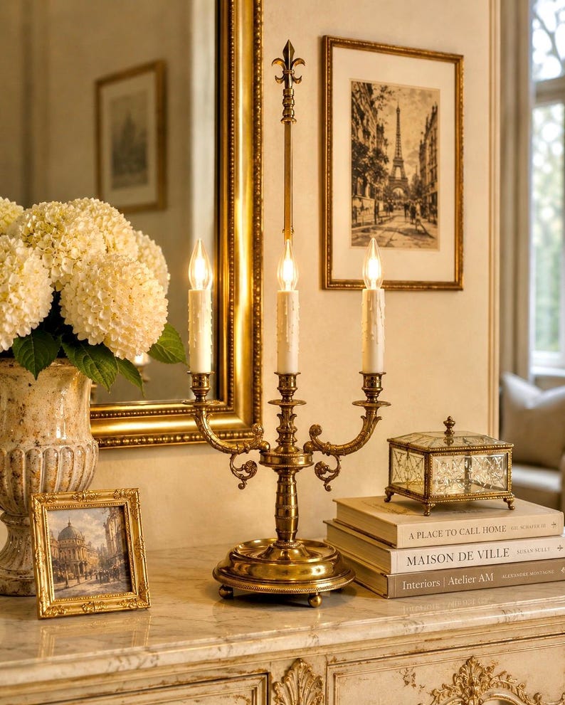 May include: A gold-toned candelabra with three candle holders, set on a marble-topped table. The candelabra is flanked by framed artwork, a decorative glass box, and a vase of white flowers. The scene evokes a classic, elegant aesthetic.