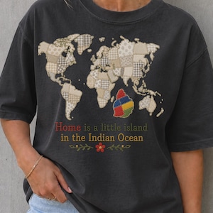 Puede incluir: Una camiseta gris carbón de manga corta con un gráfico de mapa mundial compuesto por parches de patrones beige y blancos. Debajo se destaca una pequeña isla colorida, con el texto Home is a little island in the Indian Ocean.