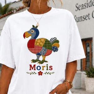 May include: A white short-sleeved t-shirt featuring a graphic of a dodo bird composed of various colourful patchwork patterns. Below the bird, the name "Moris" is printed in multicoloured letters, accompanied by a small red flower and green leaf accents.