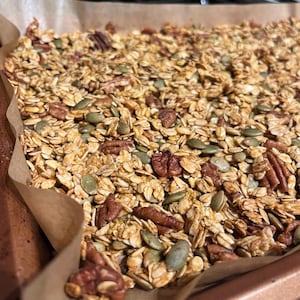 May include: A tray of homemade granola, featuring golden-brown oats, pecans, and green pumpkin seeds. The granola is baked on a sheet of baking parchment. The tray is copper-coloured.