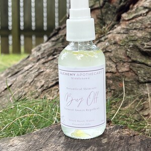 May include: A clear glass spray bottle with a white cap and label. The label reads "Bug Off, Natural Insect Repellent" from Alchemy Apothecary. The bottle contains a light green liquid and sits on a weathered wooden surface.