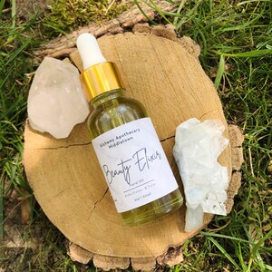Könnte beinhalten: Eine klare Glasflasche "Beauty Elixir" Gesichtsöl mit goldenem Verschluss und weißer Pipette, auf einer Holzscheibe. Das Etikett lautet "Alchemy Apothecary Middletown". Zwei klare Quarzkristalle flankieren die Flasche. Das Öl ist 60 ml.
