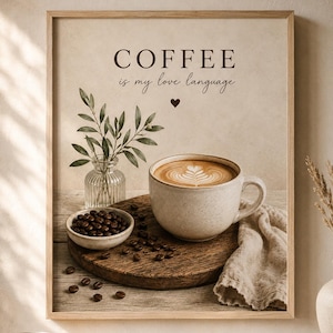 May include: A framed wall art print featuring the text COFFEE is my love language with a small heart icon. The image displays a ceramic mug with latte art, a small bowl of dark coffee beans, a glass vase with green leaves, and a linen cloth.