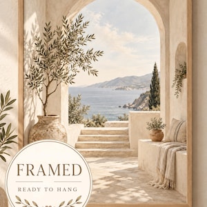 Könnte beinhalten: Ein gerahmter Kunstdruck mit einer mediterranen Küstenszene durch einen steinernen Torbogen. Das Bild zeigt eine Steinterrasse mit einem Olivenbaum in einem Tontopf, Blick auf das Meer und eine Steinbank mit einer Decke. Der Text lautet FRAMED READY TO HANG.