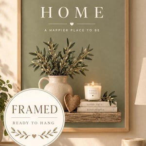 May include: A framed wall art print with a sage green background featuring the text HOME A HAPPIER PLACE TO BE. The image depicts a ceramic pitcher with olive branches, a lit candle, a wooden heart, and stacked books labelled GOOD DAYS KIND PEOPLE.