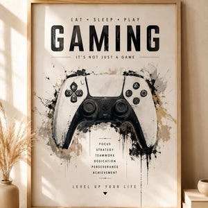 Op de afbeelding: Een ingelijste kunstdruk met een afbeelding van een wit en zwart videogame-controller met aquarelachtige spetters. De tekst luidt: EAT SLEEP PLAY GAMING, IT'S NOT JUST A GAME, FOCUS, STRATEGY, TEAMWORK, DEDICATION, PERSEVERANCE, ACHIEVEMENT, LEVEL UP YOUR LIFE.