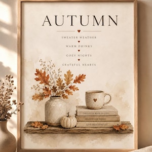 Op de afbeelding: Een ingelijste wandprint met de tekst AUTUMN, SWEATER WEATHER, WARM DRINKS, COZY NIGHTS en GRATEFUL HEARTS. De illustratie toont een vaas met oranje eikenbladeren, een kleine pompoen en een mok op een stapel van drie boeken.