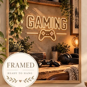 Puede incluir: Un cuadro cuadrado enmarcado que presenta un letrero de neón de tonos cálidos con la palabra GAMING sobre un icono de mando de juego. La escena muestra un escritorio con un mando, auriculares, libros y plantas, todo dentro de un marco de madera clara.