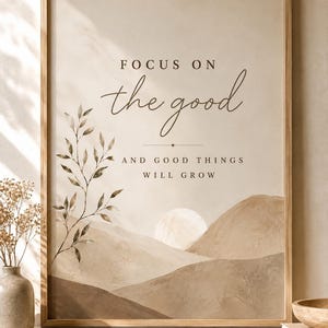 Op de afbeelding: Een ingelijste kunstdruk met een beige achtergrond en een aquarel landschap van glooiende heuvels en een zon. De tekst luidt: FOCUS ON the good AND GOOD THINGS WILL GROW. Een illustratie van een tak met bladeren staat aan de linkerkant.