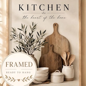 Op de afbeelding: Een ingelijste kunstdruk met de tekst KITCHEN the heart of the home. De afbeelding toont twee houten snijplanken, een keramische vaas met olijftakken, een stapel van drie kommen, een pot met deksel en houten keukengerei.