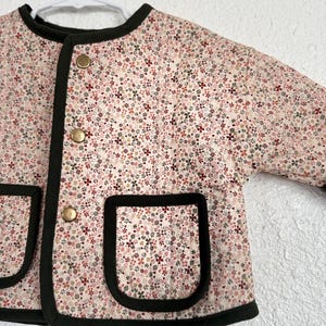 May include: A floral print jacket with a cream background and small red, green, and brown flowers. The jacket has a black trim, two front pockets, and gold-colored buttons. The jacket is hanging on a white hanger.