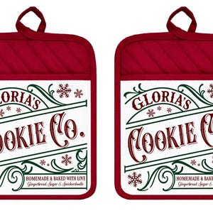 May include: Two rectangular red oven mitts with a white center panel featuring the text Gloria's Cookie Co. in dark red script and block lettering. The design includes green decorative flourishes, small snowflakes, and the text Homemade & Baked With Love.