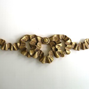 May include: A gold-colored decorative furniture applique featuring a central bow design with ruffled edges and long, flowing ribbon tails extending horizontally on both sides. The piece has a metallic finish with visible texture and relief detailing.