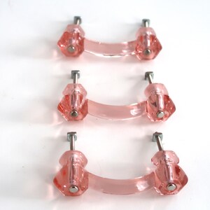 May include: Three translucent pink glass drawer pulls arranged vertically. Each pull features a curved handle connected to two faceted, hexagonal-shaped mounting points with silver-toned metal hardware extending from the back for installation.