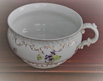 May include: White porcelain chamber pot with a floral design featuring purple flowers and gold accents. The pot has a handle on the side.