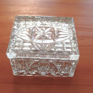 May include: A clear, square glass trinket box with a detailed, geometric pattern. The lid features a central starburst design, and the box sits on a wooden surface. The box is likely used for storing jewelry or small keepsakes.